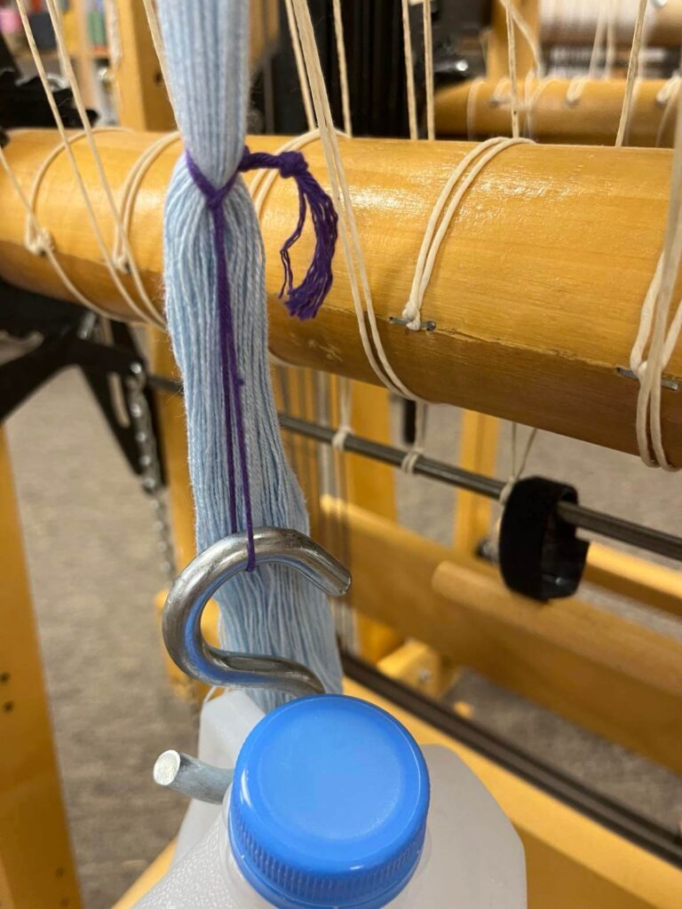 close up of an s hook attaching a jug to a loop of yarn that is attached to part of a warp
