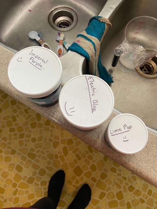 Image of three containers of dye sitting on the edge of a kitchen sink, the containers have had written labels with smiley faces on them.