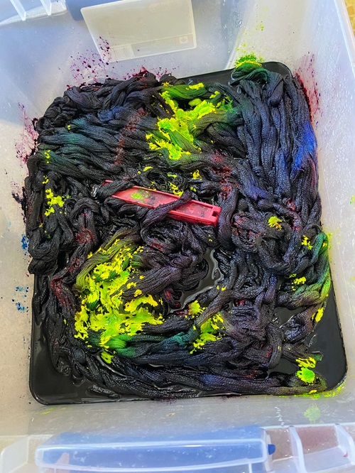 a clear bin, with chains yarn in it. The chains are in a slurry of dark dye water and there are parts of the warp that still have lime green dye powder on them