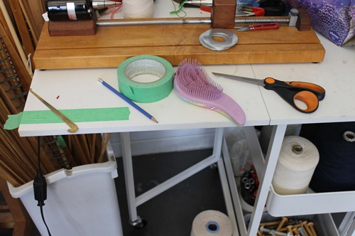 A white table with scissors, a tangle teaer brush, painters tape a sleying hook and a pencil on it