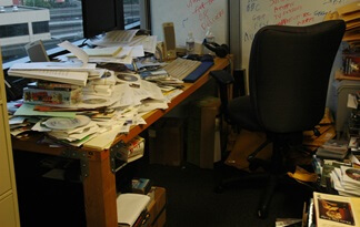 Image of a desk covered with piles of papers and a whiteboard with writing on it in the background