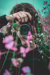 Photographer holding a camera among pink wildflowers, with a rule-of-thirds grid placing the lens near a gridline intersection.